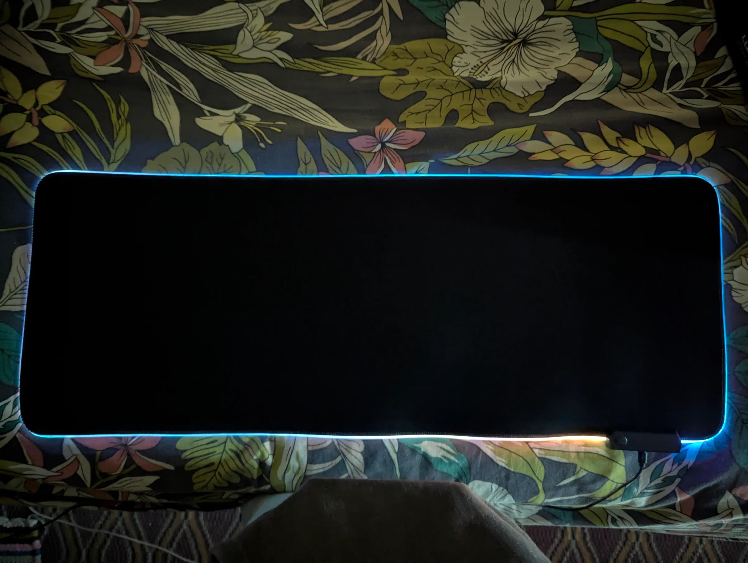 Extra-large black RGB LED desk mat glowing blue along its stitched edge, plugged in via USB on a floral bedspread